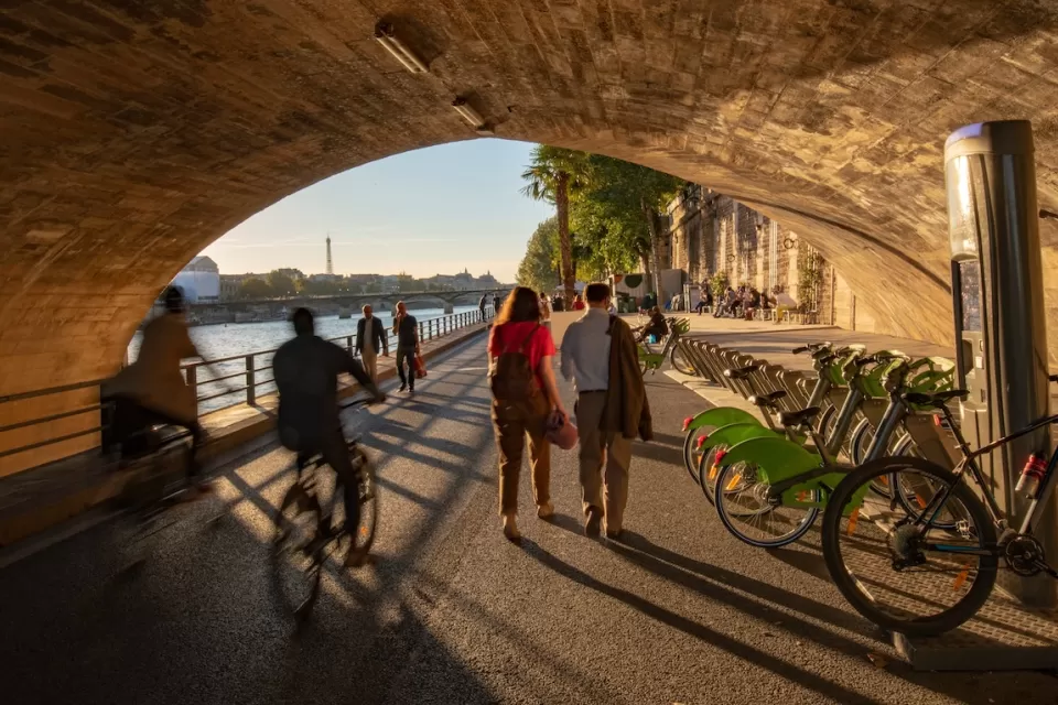 PEople on bikes, pedestrians, and a bike share station under Pont Neuf in Paris, France.